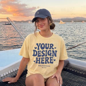 Puede incluir: Una camiseta amarilla con el texto "YOUR DESIGN HERE" en azul, con detalles de corazón. La modelo lleva una gorra de béisbol azul marino y pendientes de aro dorados. El fondo muestra una puesta de sol sobre el océano con barcos.