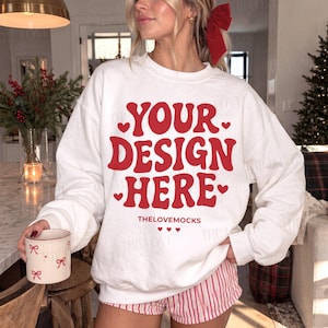 以下が含まれることがあります： 太字の波打つフォントで赤い文字「YOUR DESIGN HERE」がプリントされた白いクルーネックのスウェットシャツ。小さな赤いハートがアクセントになっています。前面のメインデザインの下には「THELOVEMOCKS」という文字と3つの小さなハートが配置されています。