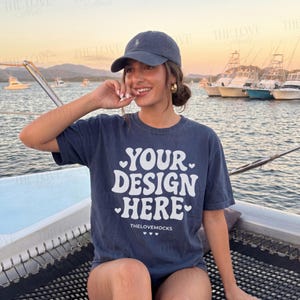 Puede incluir: Una camiseta azul marino con el texto "YOUR DESIGN HERE" en blanco, y una gorra de béisbol azul marino. La camiseta la lleva una persona en un barco, con barcos y agua al fondo.