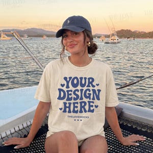 May include: A cream-colored t-shirt with the text "YOUR DESIGN HERE" in blue, with heart accents. The model is wearing a navy blue baseball cap and gold hoop earrings. The background shows a body of water and boats.