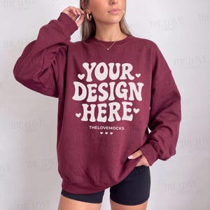 May include: A maroon long-sleeve crewneck sweatshirt featuring the white text "YOUR DESIGN HERE" in a bold, wavy font, accented by small white hearts. The text "THELOVEMOCKS" is printed in smaller white letters below the main design.