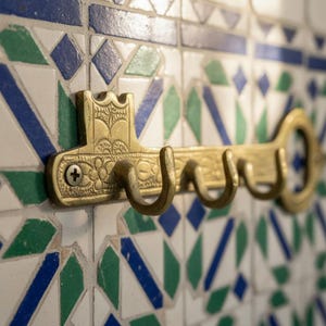 Op de afbeelding: Een messing sleutelvormige wandhaak met drie haken, gemonteerd op een betegelde muur met patronen. De haak heeft sierlijke details en een decoratieve sleutelkop. De tegels hebben blauwe, groene en witte geometrische ontwerpen.