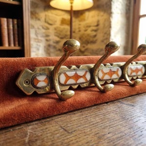 May include: A vintage brass coat rack with three curved hooks. The rack has decorative rectangular inlays with white and orange starburst patterns. It is mounted on a rust-coloured velvet backing.