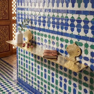 Puede incluir: Estante de metal dorado texturizado, montado en una pared con azulejos estampados azules, verdes y blancos. Contiene un jarrón blanco con vegetación, un diario de cuero marrón y detalles dorados. La estantería mide unos 60 cm de largo.