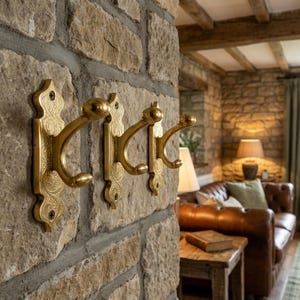 May include: Three ornate brass wall hooks mounted on a textured stone wall. Each hook has a decorative backplate and a curved hook design. The hooks are a warm gold color, adding a touch of elegance to the rustic setting.
