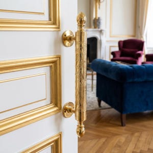 Könnte beinhalten: Verzierter goldener Türgriff mit detailliertem Blumen- und Rankenmuster. Der Griff ist an einer weißen Tür mit goldenen Zierleisten befestigt. Das Design umfasst dekorative obere und untere Zierknöpfe. Der Hintergrund zeigt einen Raum mit einem blauen Sofa und einem Kamin.