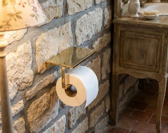 May include: A gold-colored metal toilet paper holder mounted on a stone wall. The holder has a small shelf above the roll of white toilet paper. A wooden lamp and a vintage vanity are in the background.