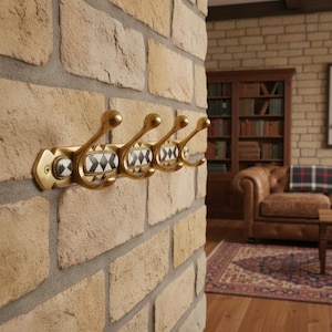 Op de afbeelding: Een messingkleurige wandkapstok met vijf gebogen haken. Elke haak is versierd met een decoratief paneel met een zwart-wit geometrisch patroon. De kapstok is bevestigd aan een bakstenen muur, wat wijst op een klassieke of vintage stijl.