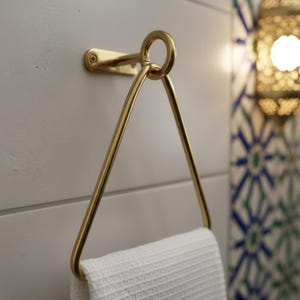 May include: A gold-colored metal towel rack mounted on a white wall. The rack has a triangular design with a circular top and holds a white textured towel. The bathroom fixture is a modern design.