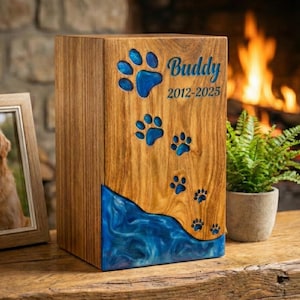 May include: Wooden pet urn with blue paw prints and a blue resin wave design. The name "Buddy" and dates 2012-2025 are inscribed on the front. A framed photo of a golden retriever is on the left.