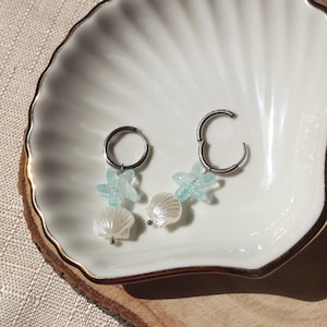 Könnte beinhalten: Silberne Creolen mit maritimen Anhängern. Jeder Ohrring hat einen hellblauen Seestern und einen weißen Muschelanhänger. Die Ohrringe werden auf einer weißen muschelförmigen Schale mit goldenem Rand präsentiert.