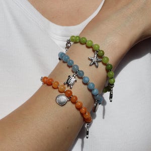 Könnte beinhalten: Drei Perlenarmbänder mit glatten, runden Perlen in den Farben Limettengrün, Hellblau und Orange. Jedes Armband ist mit einem silberfarbenen Metallanhänger in Form eines Seesterns, einer Meeresschildkröte oder einer Muschel versehen.