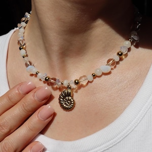 Könnte beinhalten: Eine Perlenkette mit einem zentralen Anhänger in Form eines goldenen Ammoniten. Die Kette besteht aus abwechselnd klaren, irisierenden und undurchsichtigen weißen Perlen, die mit kleinen, polierten, goldfarbenen Metallkugeln durchsetzt sind.