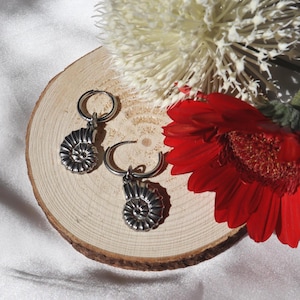 Könnte beinhalten: Silberne Creolen mit detailliertem Ammoniten-Fossil-Design. Die Ohrringe werden auf einer Holzscheibe mit einer roten Gerbera und einer weißen Blume im Hintergrund präsentiert. Einzigartiger Schmuck.