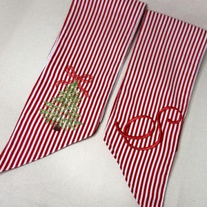 Peut inclure: Deux bandes de tissu avec un motif à rayures verticales rouges et blanches. L'une présente un sapin de Noël vert brodé surmonté d'un nœud rouge. L'autre arbore une lettre S cursive rouge brodée sur le tissu rayé.