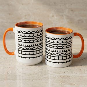 Puede incluir: Dos tazas de cerámica blanca con asas y bordes interiores de color naranja, cada una llena de una bebida oscura humeante. El exterior de ambas tazas presenta un patrón geométrico negro repetitivo de líneas, triángulos y semicírculos.
