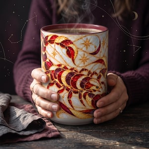 May include: A ceramic mug featuring a stained-glass style illustration of a scorpion in shades of deep red, burgundy, and cream. The design includes gold-toned outlines of stars and celestial patterns against a light background.