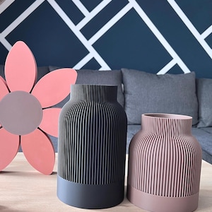Könnte beinhalten: Zwei moderne Vasen mit vertikalen Rippenmustern. Eine ist schwarz mit dunkelgrauem Sockel, die andere hellrosa mit mauvefarbenem Sockel. Links befindet sich eine große rosa Blumendekoration.