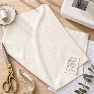 May include: Cream-colored fabric with a subtle dotted pattern, alongside sewing tools. Gold scissors, a measuring tape, and a spool of thread are visible. A tag indicates the fabric is 100% cotton with care instructions.