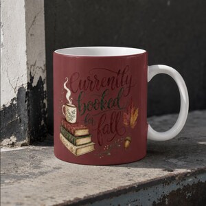 May include: A dark red ceramic mug with a white interior and handle. The mug features a graphic of three stacked books, a small mug with steam, an acorn, and a leaf. The text "Currently booked for fall" is printed in a dark cursive script.