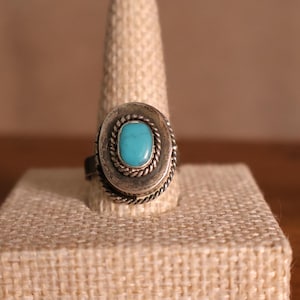 May include: A silver ring with an oval turquoise stone set in a decorative frame. The ring is displayed on a beige linen ring holder, showcasing the jewelry's design and color. The ring's band is visible, indicating its size and style.