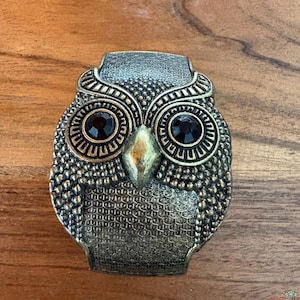 May include: A vintage-style owl-shaped metal clasp with a bronze finish. The owl has black gem eyes and a textured surface. The clasp is set against a wooden background.