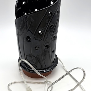May include: Black ceramic essential oil diffuser with a glossy finish. The diffuser has a unique, curved design with decorative holes and a brown base. It includes a clear power cord with a plug.