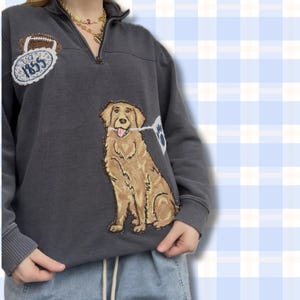 May include: Charcoal gray pullover sweatshirt with a quarter-zip neckline. Features an embroidered golden retriever holding a paw print flag. A football and the text "1855" are embroidered on the left chest. The sweatshirt has ribbed cuffs and hem.