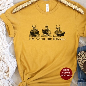 Peut inclure: Un t-shirt jaune moutarde avec un graphique de trois squelettes lisant des livres. Le texte "I'm with the banned" est imprim&eacute; sous les squelettes. Le t-shirt a un cercle rouge avec le texte "More Colors Available" en blanc.