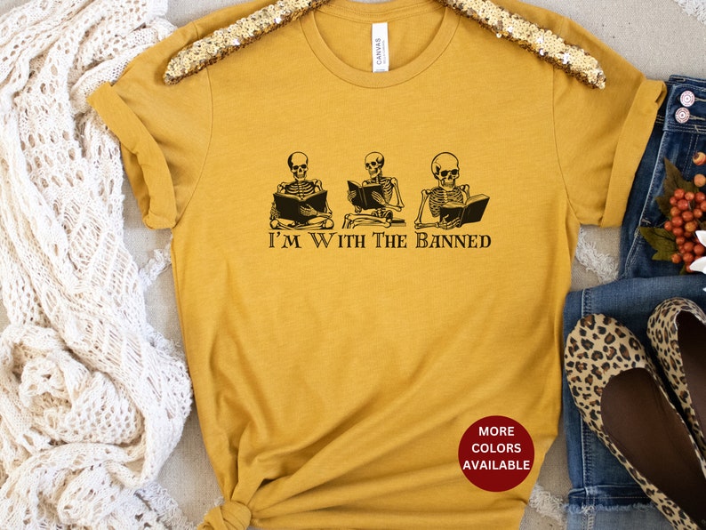 Peut inclure: Un t-shirt jaune moutarde avec un graphique de trois squelettes lisant des livres. Le texte "I'm with the banned" est imprim&eacute; sous les squelettes. Le t-shirt a un cercle rouge avec le texte "More Colors Available" en blanc.