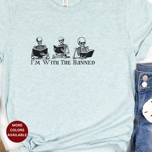 Peut inclure: Un t-shirt bleu clair avec un graphique de trois squelettes lisant des livres. Le texte "I'm With The Banned" est imprim&eacute; sous les squelettes.