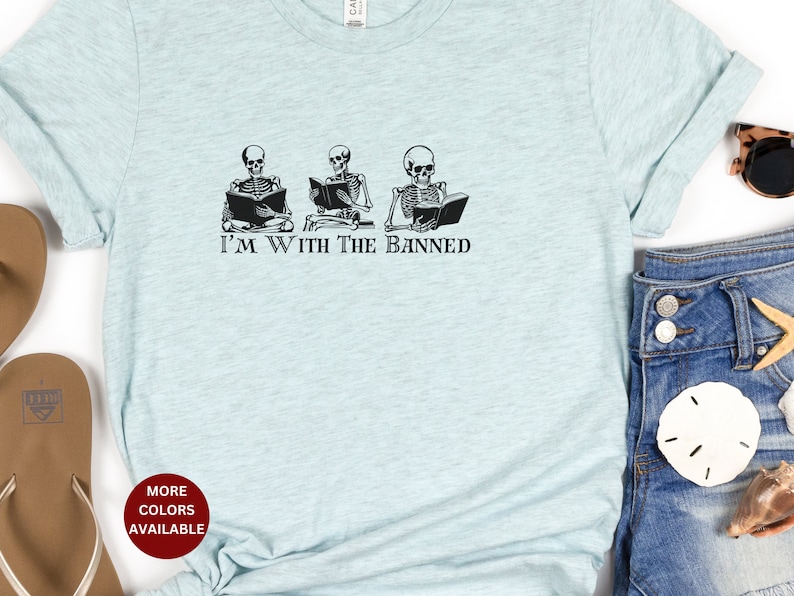 Peut inclure: Un t-shirt bleu clair avec un graphique de trois squelettes lisant des livres. Le texte "I'm With The Banned" est imprim&eacute; sous les squelettes.
