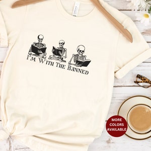 Peut inclure: Un t-shirt blanc avec un graphique de trois squelettes lisant des livres. Le texte "I'm with the banned" est imprim&eacute; sous les squelettes.