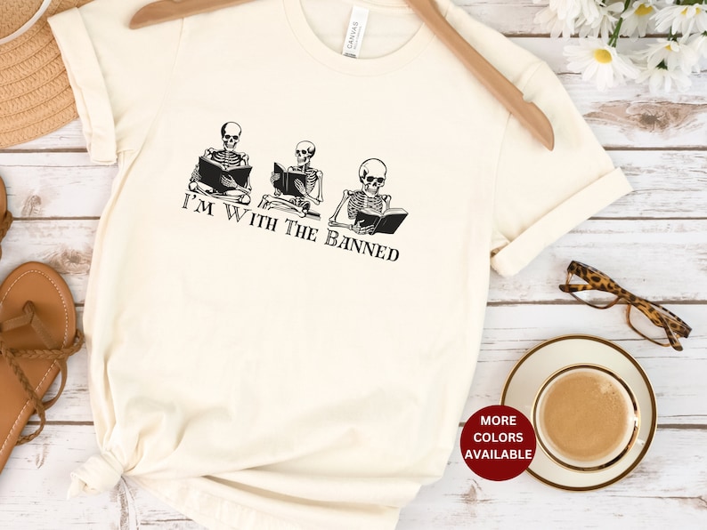 Peut inclure: Un t-shirt blanc avec un graphique de trois squelettes lisant des livres. Le texte "I'm with the banned" est imprim&eacute; sous les squelettes.
