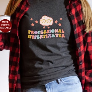 May include: A dark gray t-shirt with a cartoon brain wearing glasses and the text "PROFESSIONAL HYPERFIXATER" in rainbow colors.