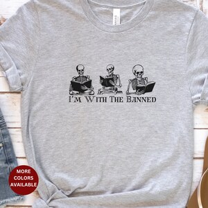Peut inclure: T-shirt gris avec un motif de trois squelettes lisant des livres. Le texte "I'm With The Banned" est imprim&eacute; sous les squelettes.