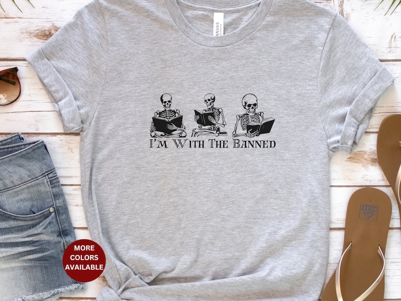 Peut inclure: T-shirt gris avec un motif de trois squelettes lisant des livres. Le texte "I'm With The Banned" est imprim&eacute; sous les squelettes.