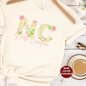 Pu&ograve; includere: Una maglietta bianca con un design floreale che compone la scritta "NC" per la Carolina del Nord. La maglietta ha un nodo sul fondo ed &egrave; appoggiata su una superficie di legno con una tazza di caff&egrave;, occhiali e un cappello di paglia.