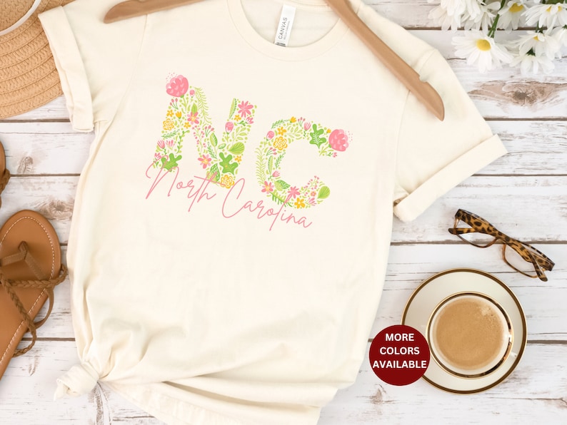 Pu&ograve; includere: Una maglietta bianca con un design floreale che compone la scritta "NC" per la Carolina del Nord. La maglietta ha un nodo sul fondo ed &egrave; appoggiata su una superficie di legno con una tazza di caff&egrave;, occhiali e un cappello di paglia.