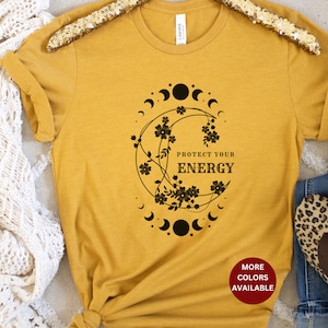 May include: A mustard yellow t-shirt with a black graphic of a crescent moon with flowers and the text "Protect Your Energy". The shirt has a rolled up sleeve and a tag that says "More Colors Available".