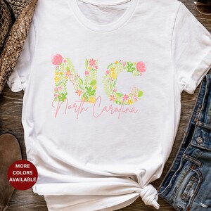 Pu&ograve; includere: Maglietta bianca con un design floreale che compone la scritta "NC" e le parole "North Carolina" sotto.