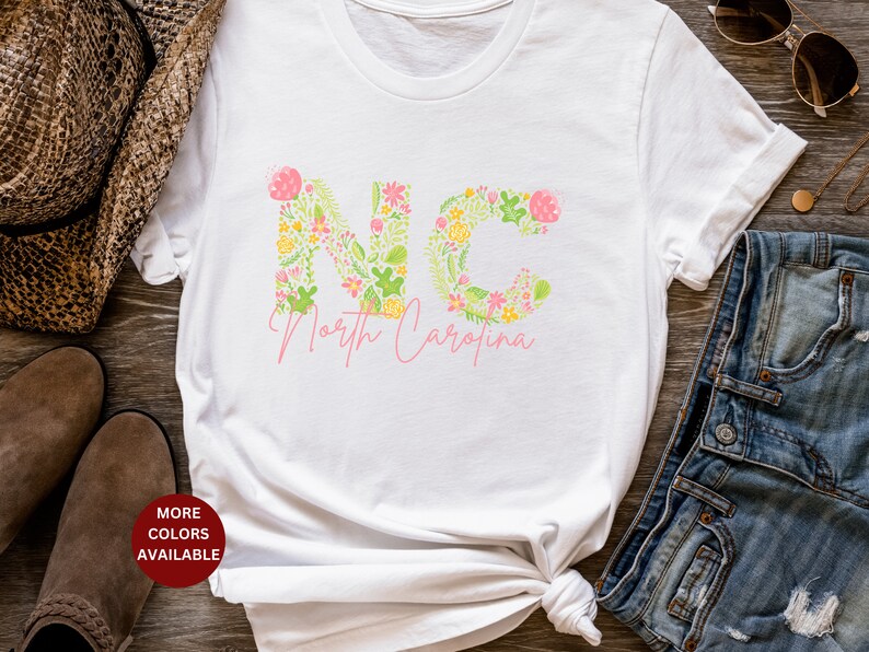 Pu&ograve; includere: Maglietta bianca con un design floreale che compone la scritta "NC" e le parole "North Carolina" sotto.