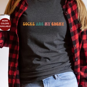 May include: A dark gray t-shirt with the text "Socks are my enemy" in colorful letters. The shirt is being worn by a person wearing a red and black plaid shirt.