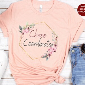 May include: Pink t-shirt with a gold hexagonal frame and floral design. The text "Chaos Coordinator" is inside the frame.