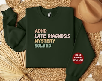 May include: A dark green sweatshirt with the text "ADHD Late Diagnosis Mystery Solved" in a colorful retro font. The text is in shades of pink, orange, and yellow. The sweatshirt has a crew neck and long sleeves. The text "More Colors Available" is on a red circle on the right sleeve.