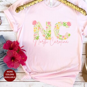 Pu&ograve; includere: Maglietta rosa con le lettere NC in stampa floreale e il testo "North Carolina" sotto.