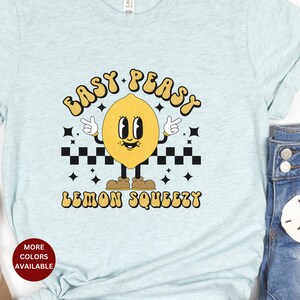 K&ouml;nnte beinhalten: Ein hellblaues T-Shirt mit einer gelben Cartoon-Zitrone, die braune Schuhe tr&auml;gt und den Text "Easy Peasy Lemon Squeezy" in einer Retro-Schrift.
