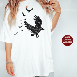 May include: White t-shirt with a black silhouette of a raven with stars inside its wings. The raven is flying with other smaller ravens flying behind it. The text "MORE COLORS AVAILABLE" is in a red circle on the bottom right of the image.