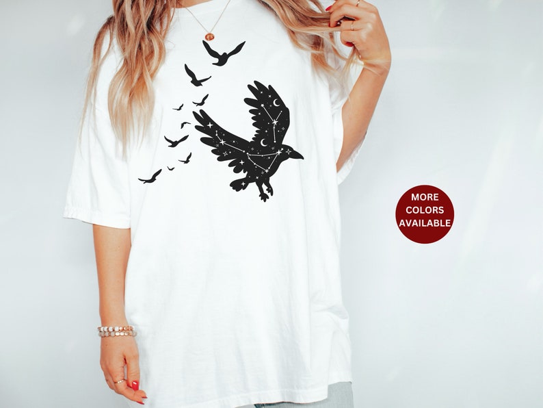 May include: White t-shirt with a black silhouette of a raven with stars inside its wings. The raven is flying with other smaller ravens flying behind it. The text "MORE COLORS AVAILABLE" is in a red circle on the bottom right of the image.