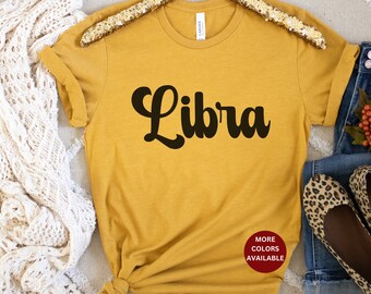 May include: A mustard yellow t-shirt with the word "Libra" in black script. The shirt has a crew neck and short sleeves. The text "More Colors Available" is in red on a white circle.